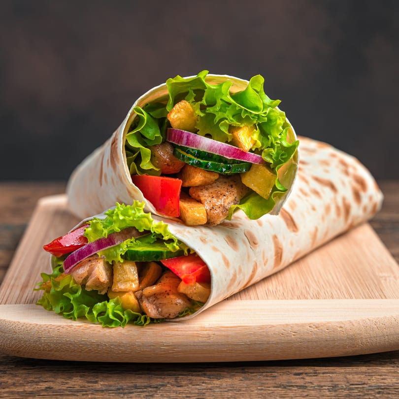 Two Chi Chi's Tacoria wraps filled with chicken, lettuce, red onion, and other vegetables on a wooden board.