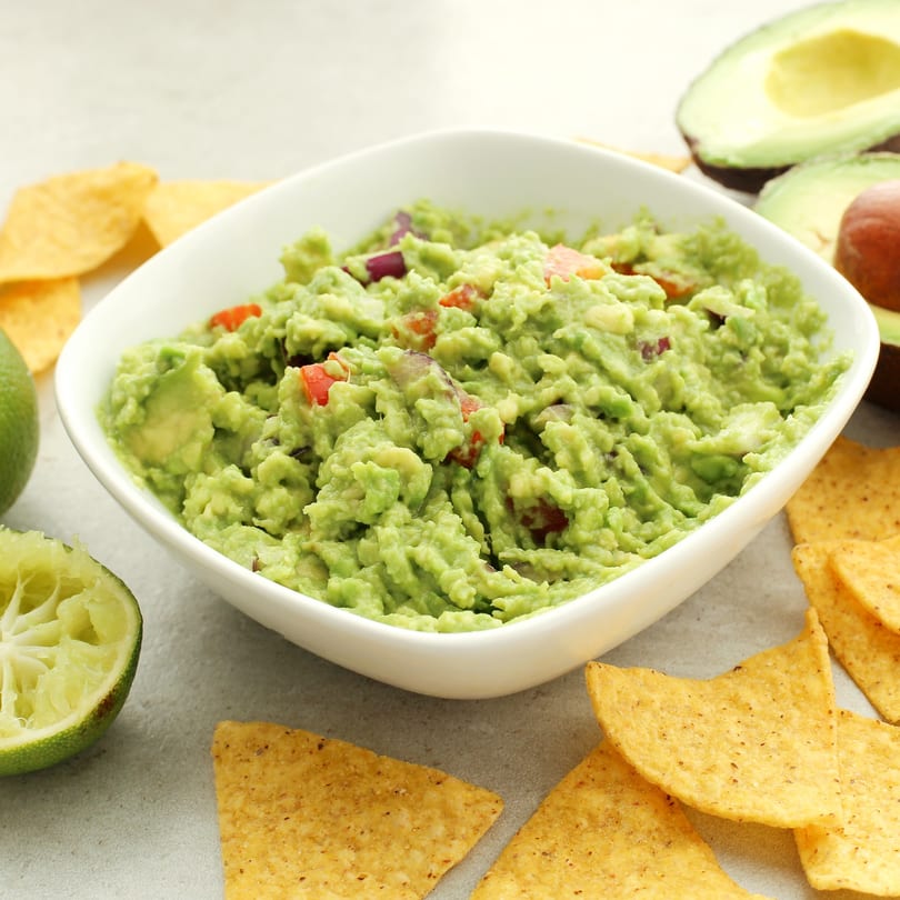 A white bowl of Chi Chi's guacamole with tortilla chips, limes, and avocados on a light surface.