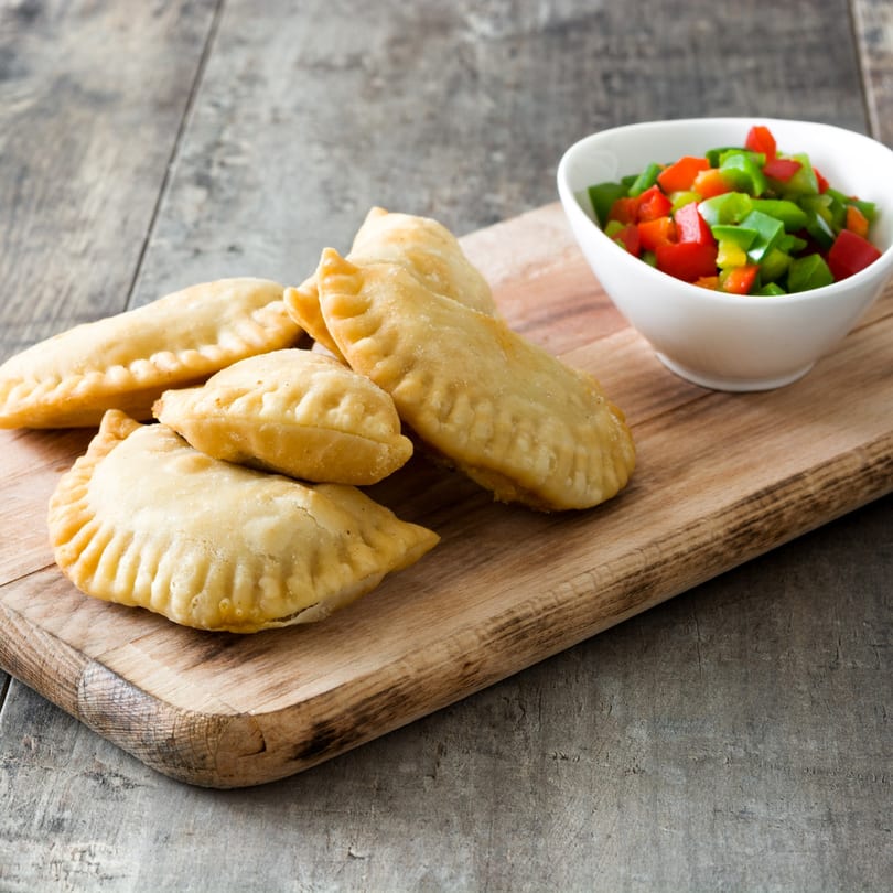 Golden-brown empanadas on a wooden board next to a white bowl of diced red and green peppers.