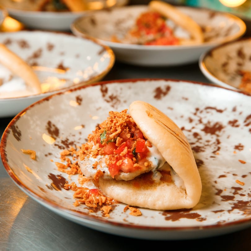 A bao bun taco with chicken, white sauce, diced tomatoes, and crispy fried onions on a patterned plate.